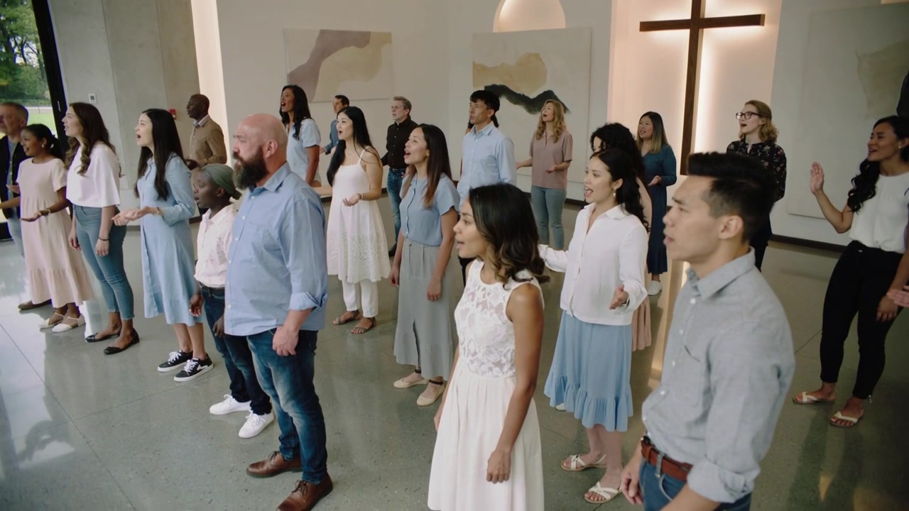 Coro cantando en la iglesia, vistiendo colores pastel mientras la música comienza, levantando las manos en adoración cerca de una cruz iluminada.