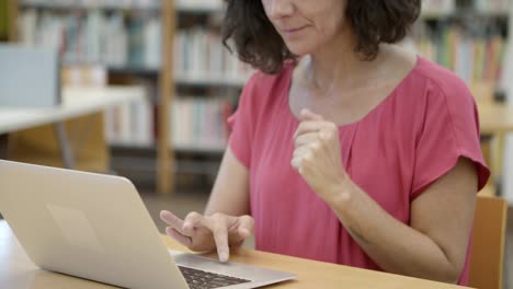 serious student sitting at table in library and working with laptop