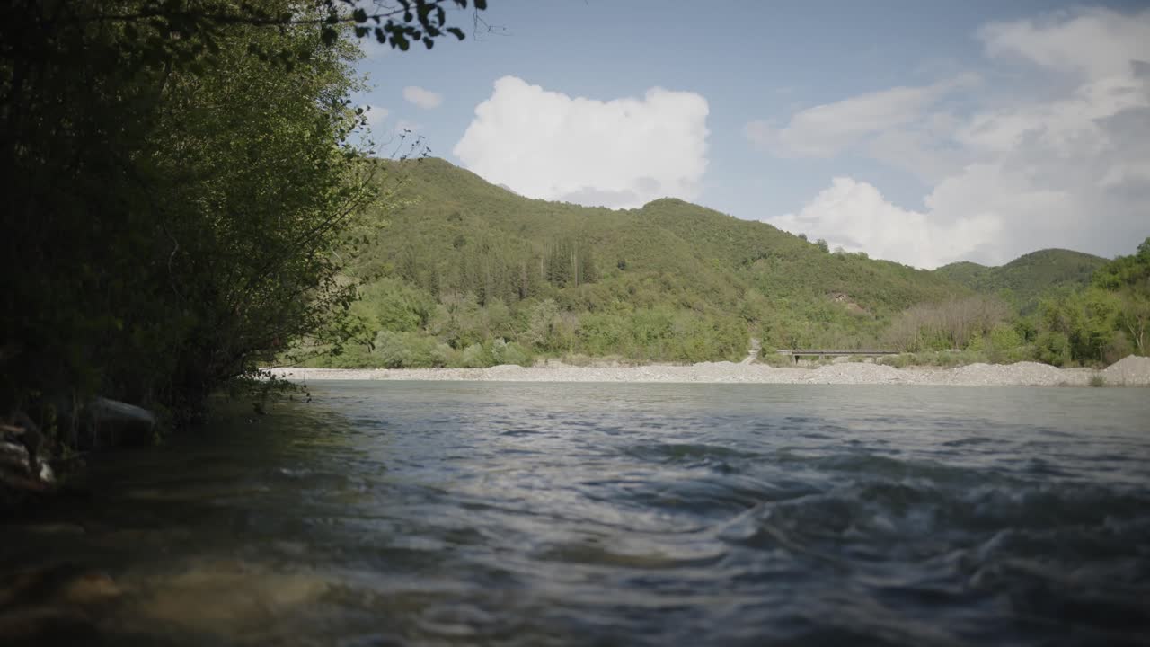 Low-angle shot from the river’s edge showing clear flowing water, distant forested hills, and a bright sky with puffy clouds.