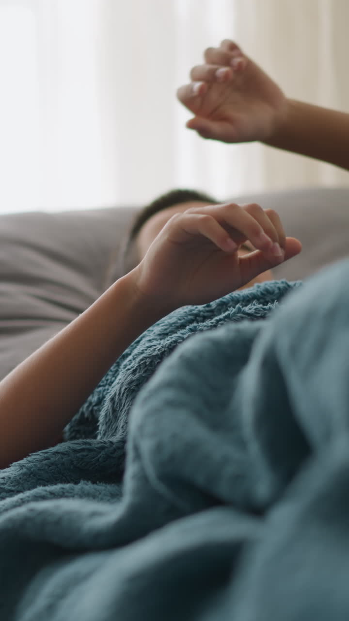 Sick child lying on couch covered with blanket, coughing and resting hand on head, tissue nearby with blurred cup and tissue on table