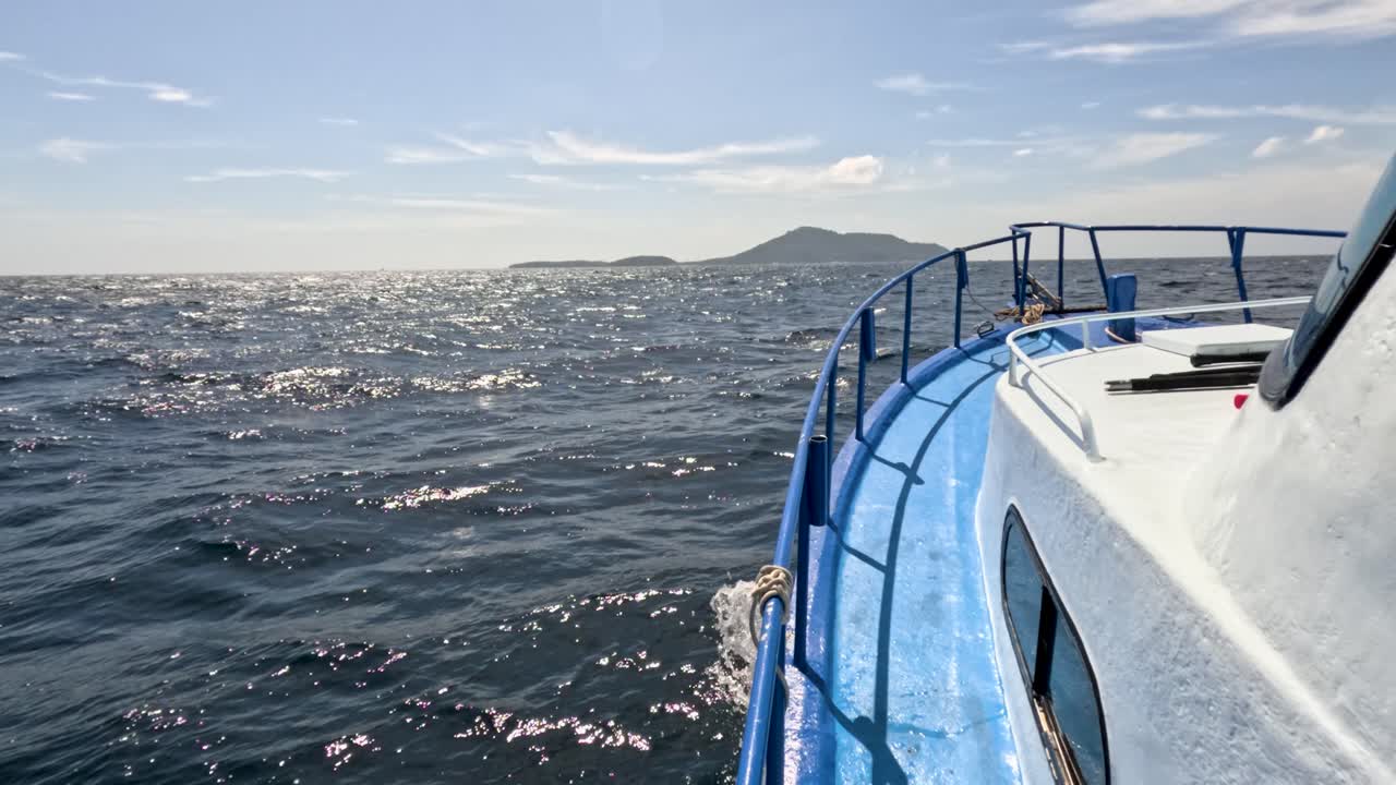 Blue boat moves across sparkling sea toward distant island, daylight, wide shot, steady camera