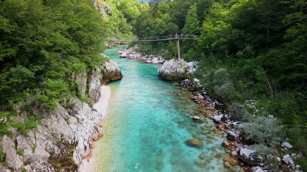 Aerial drone view of the turquoise river running between rocky banks with a small suspension bridge crossing it downstream, Soca Valley, Slovenia