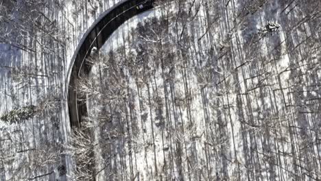 Winter-landscape-with-driving-cars-at-a-snow-framed-road,-and-look-up-to-a-sunny-landscape,-captured-by-a-drone