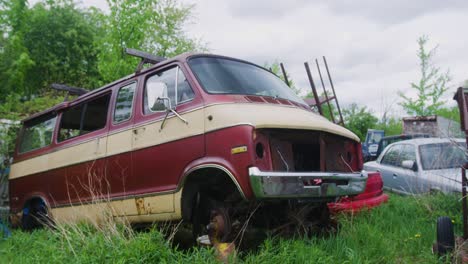 semi-orbiting shot of an abandoned cargo van without headlights or tires resting next to some other cars in an overgrown forest