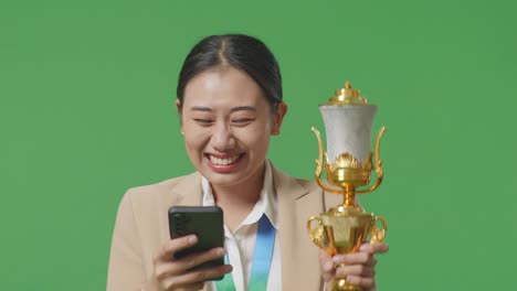 close up of asian business woman in a suit with a gold medal and trophy looking at smartphone then screaming goal celebrating on green screen background in the studio