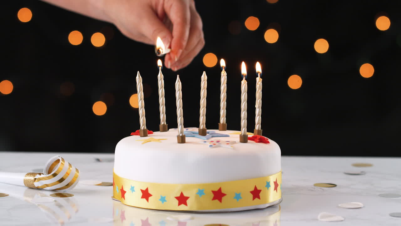 cerca de una mano usando un fósforo para encender siete velas en un pastel de cumpleaños blanco y decorado, un soplador de fiesta a su lado, luces bokeh en el fondo