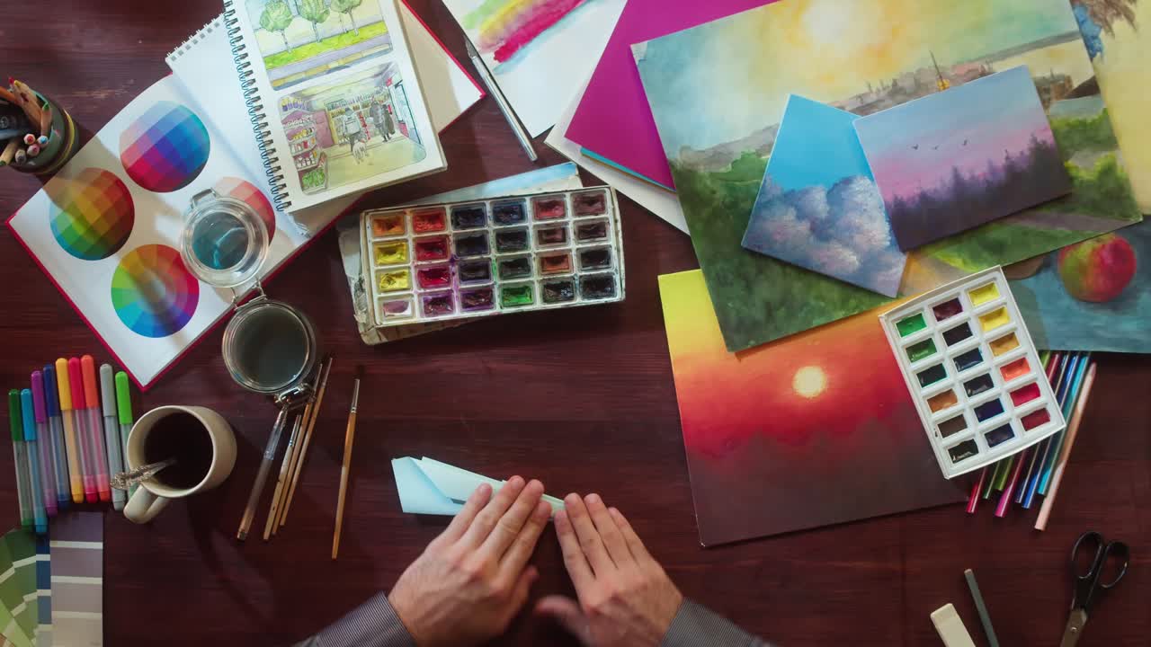 Hands Folding a Paper Airplane on an Artist's Desk with Art Supplies