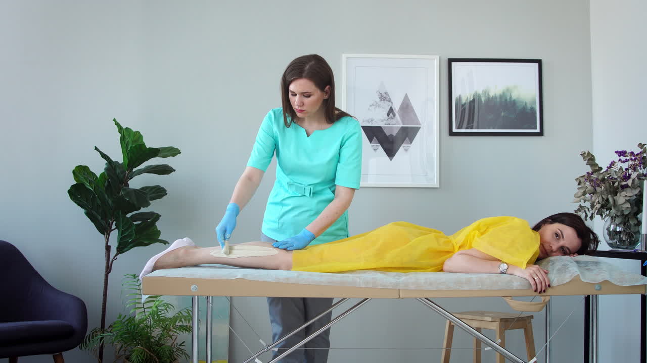 la chica habla por teléfono y sonríe durante la depilación de las piernas en el salón de belleza. depilación de las piernas con azúcar. maestro de depilación