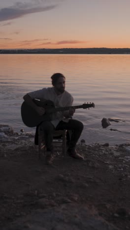 man playing guitar by the water at sunset