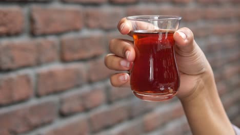 mano sosteniendo un vaso de té turco frente a una pared de ladrillo