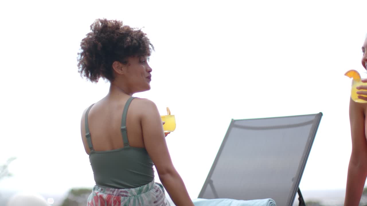 Diverse female friends in swimwear relaxing poolside, one entering carrying cocktail, sparking chat