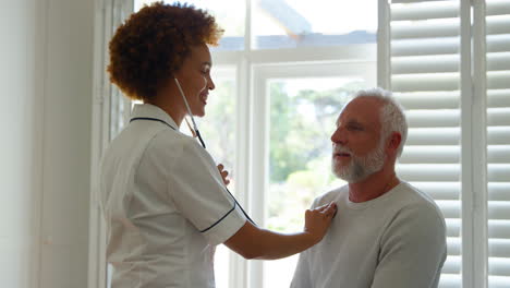 female nurse wearing uniform listening to senior male patient's chest in private hospital room