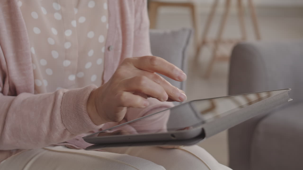 Steadicam of cropped unrecognizable person wearing pink outfit, scrolling on screen of tablet computer, sitting indoors