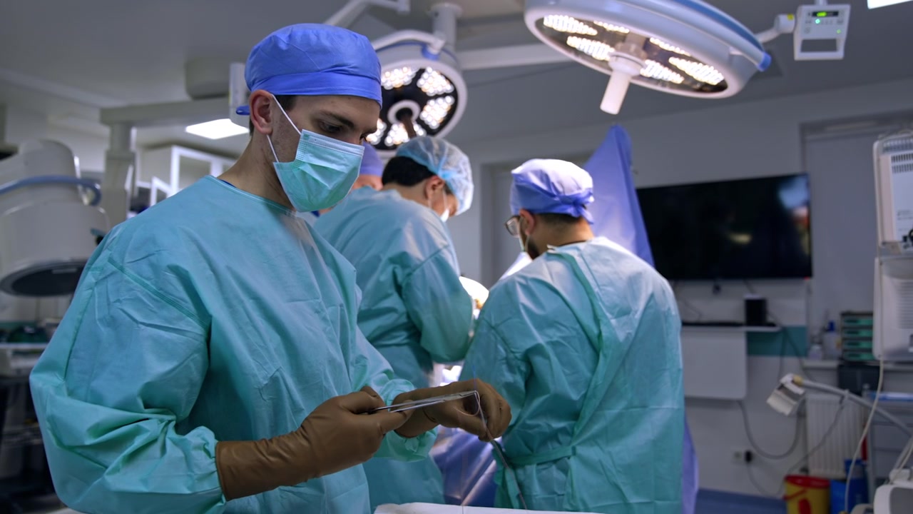 Young mid-aged male assistant arranges tools at the surgical stand and preparing instruments and materials for the doctors. Three surgeons perform trauma surgery at backdrop.