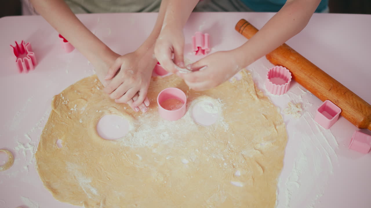 Kids pressing pink shape cutters into dough while another rubs flour on dough and quickly drops flour to cut another shape, with flour, dough, and colorful tools