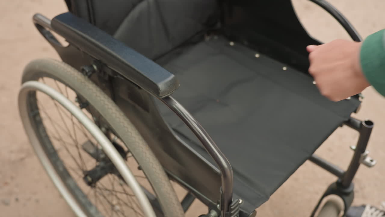 CloseUp Of Folded Wheelchair Being Placed Into Car Trunk, Compact Frame And Wheels Shown, Careful Hands Maneuvering Hinge And Release Mechanism, Outdoor Parking Area Indicating Transition