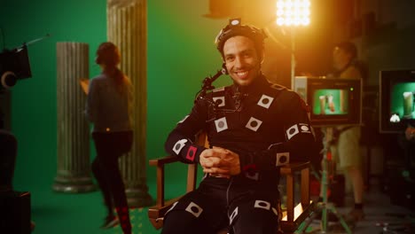 hombre guapo y sonriente con traje de captura de movimiento y equipo de cabeza teniendo descanso para almorzar, sentado en una silla, mirando a la cámara. estudio película de alto presupuesto. en el estudio de cine período de disfraces drama set de cine