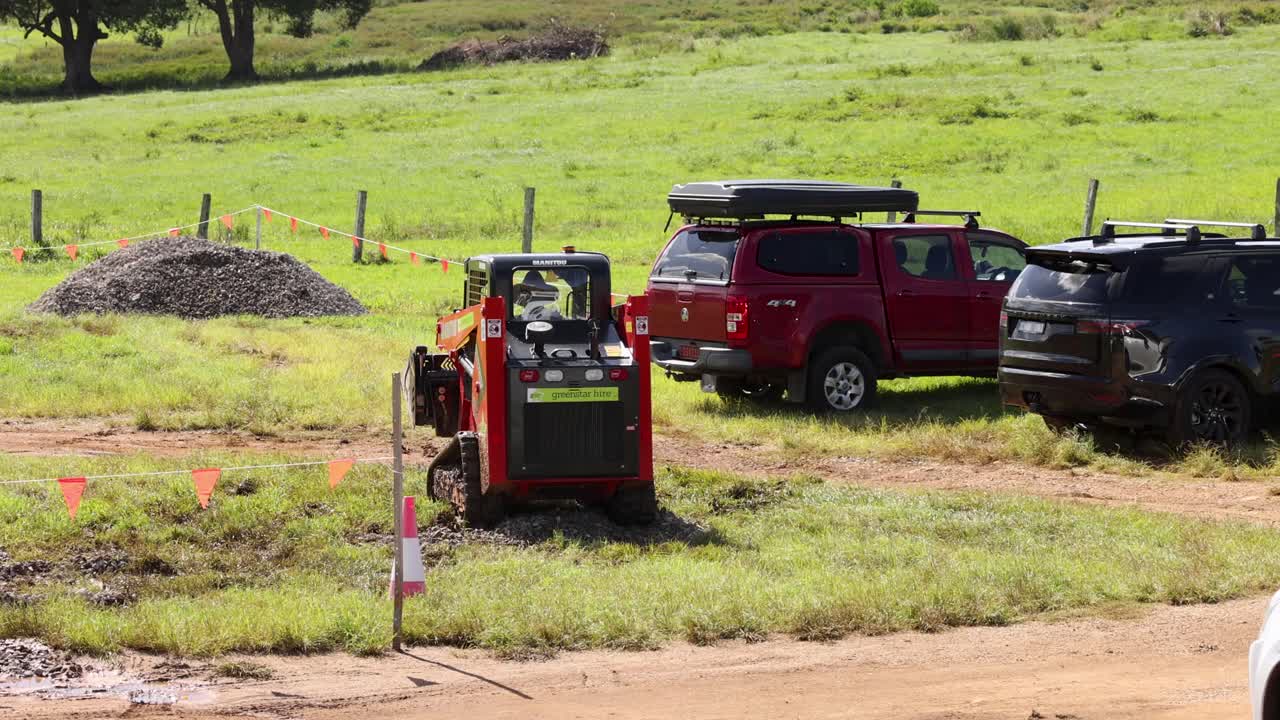 A mini skid steer loader maneuvers on a sunny farm, surrounded by vehicles and green fields, showcasing agricultural machinery in action