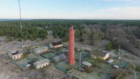 Aerial-establishing-view-of-red-colored-Akmenrags-lighthouse,-Baltic-sea-coastline,-Latvia,-white-sand-beach,-calm-sea,-sunny-day-with-clouds,-wide-drone-shot-moving-backward