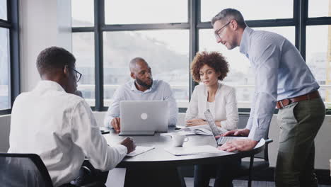 Middle-aged-white-businessman-stands-addressing-corporate-colleagues-at-a-table-in-a-meeting-room