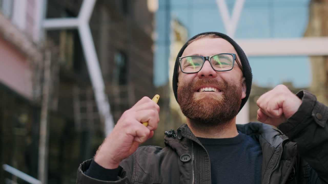 A man with a beard and glasses smiles broadly while holding a smartphone outdoors