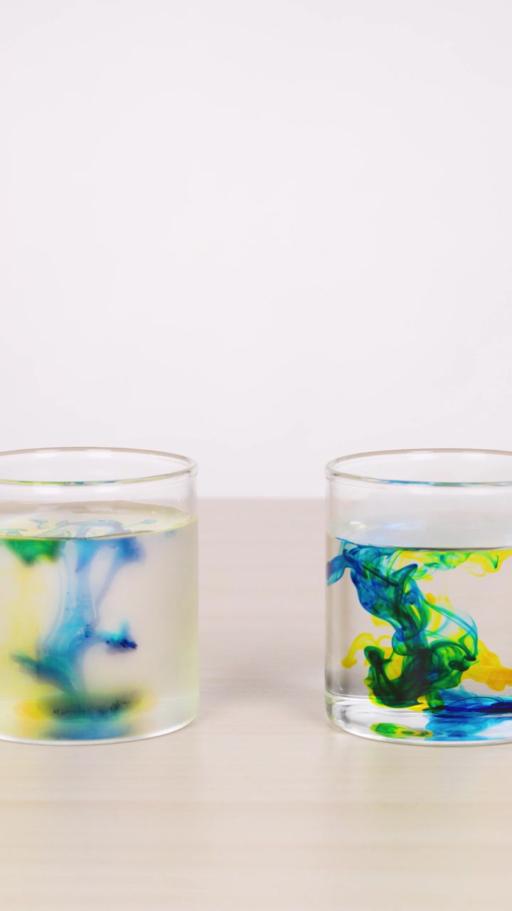 Gocce d'acqua colorate si disperdono in bicchieri trasparenti, creando motivi a spirale sotto una luce brillante e uniforme.