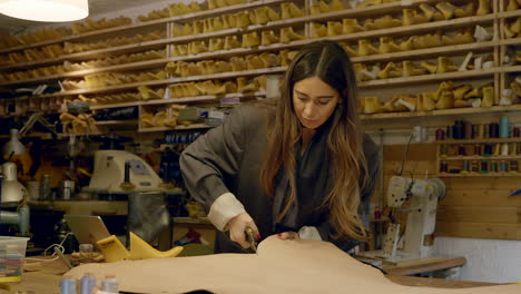 female craftsperson cutting leather pattern with shears making shoes in small business workshop