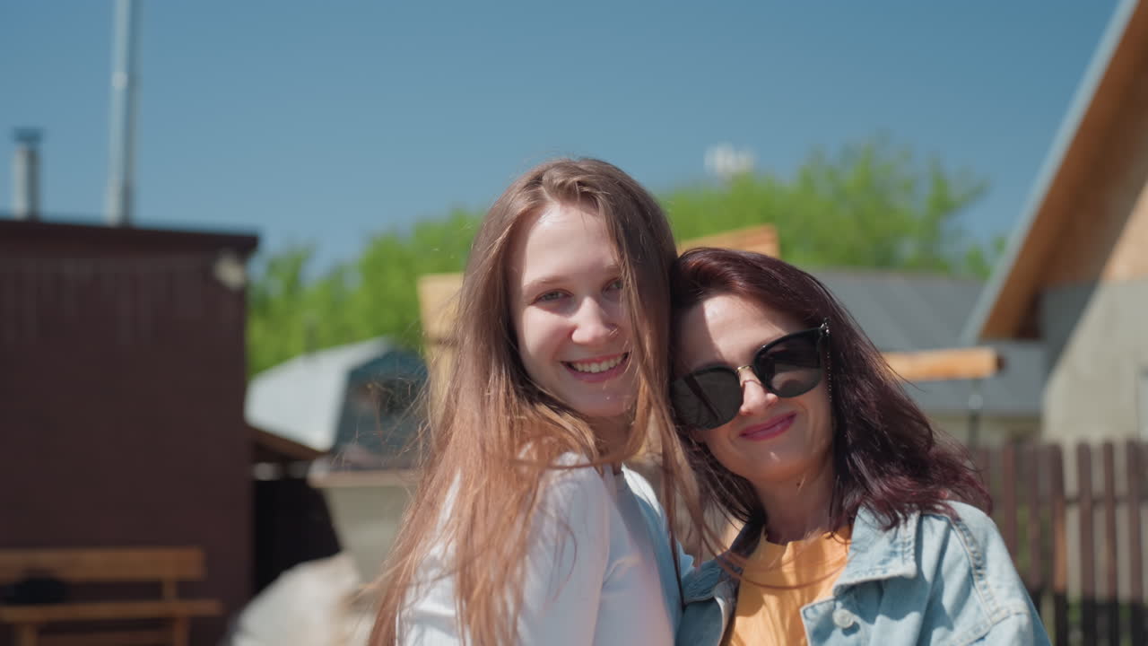 Two sisters embrace each other warmly with joyful smiles in sunlit backyard, standing in front of wooden house and garden while another girl is seen blurred lying on wooden swing in background