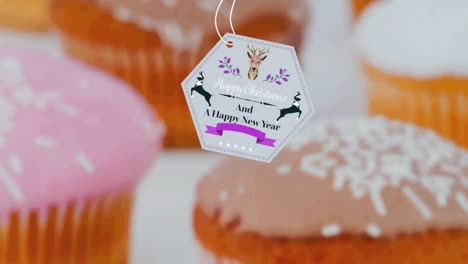 Animation-of-christmas-greetings-on-tag-over-cupcakes-on-white-background