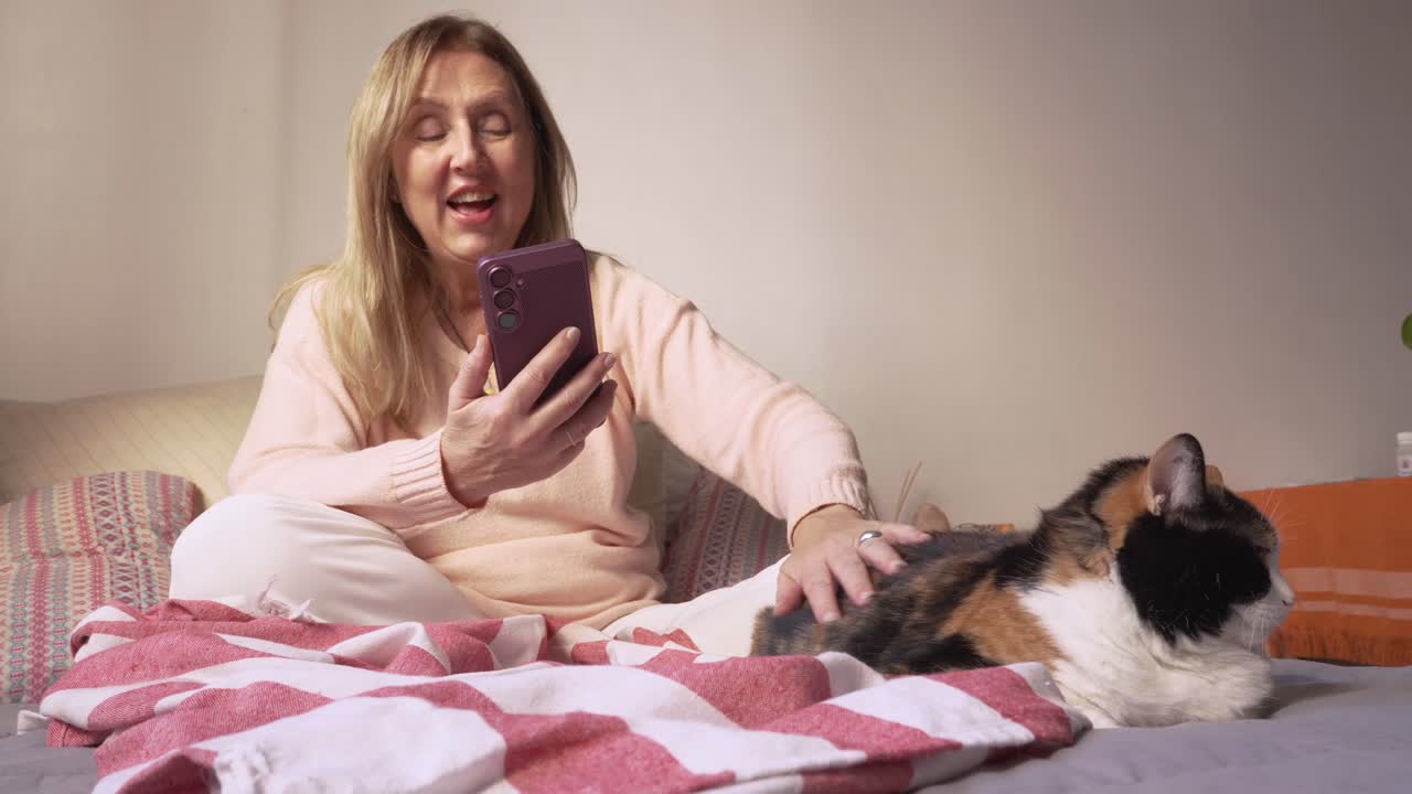 A relaxed woman enjoys a video call on smartphone while caress her calico cat comfortably on bed