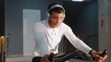 athlete in white activewear cycles on stationary bike wearing headphones under natural light streaming through gym windows, focused posture and modern equipment visible in subtle blurred background