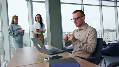 stressed angry male employee sitting at desk tears a piece of paper. women standing at backdrop look at male colleague. blonde women remarks about man's behavior but he throws the papers around.