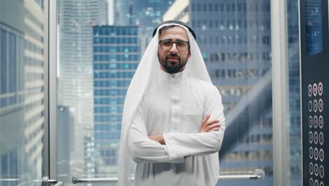 portrait of a successful happy muslim businessman in traditional white kandura wearing glasses riding glass elevator to office in modern business center. saudi, emirati, arab businessman concept.