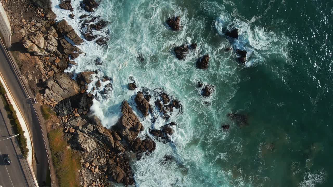 Vista aerea dall'alto di onde oceaniche che si infrangono su una costa rocciosa, lungo una strada costiera illuminata dal sole.