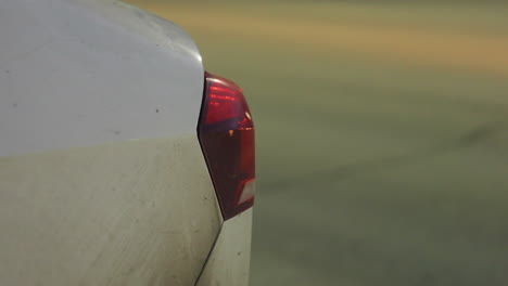 close up of vehicle tail light showing rear corner of parked car, illuminated by surrounding street lights, with blurred background of road, moving cars, and pedestrians during night time