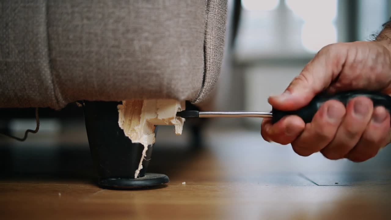 Rechte Hand führt Schraubendreherspitze ein, um zerrissene Polsterung aus der Couch zu Hause für die Reparatur zu hebeln, schwarzes Bein.