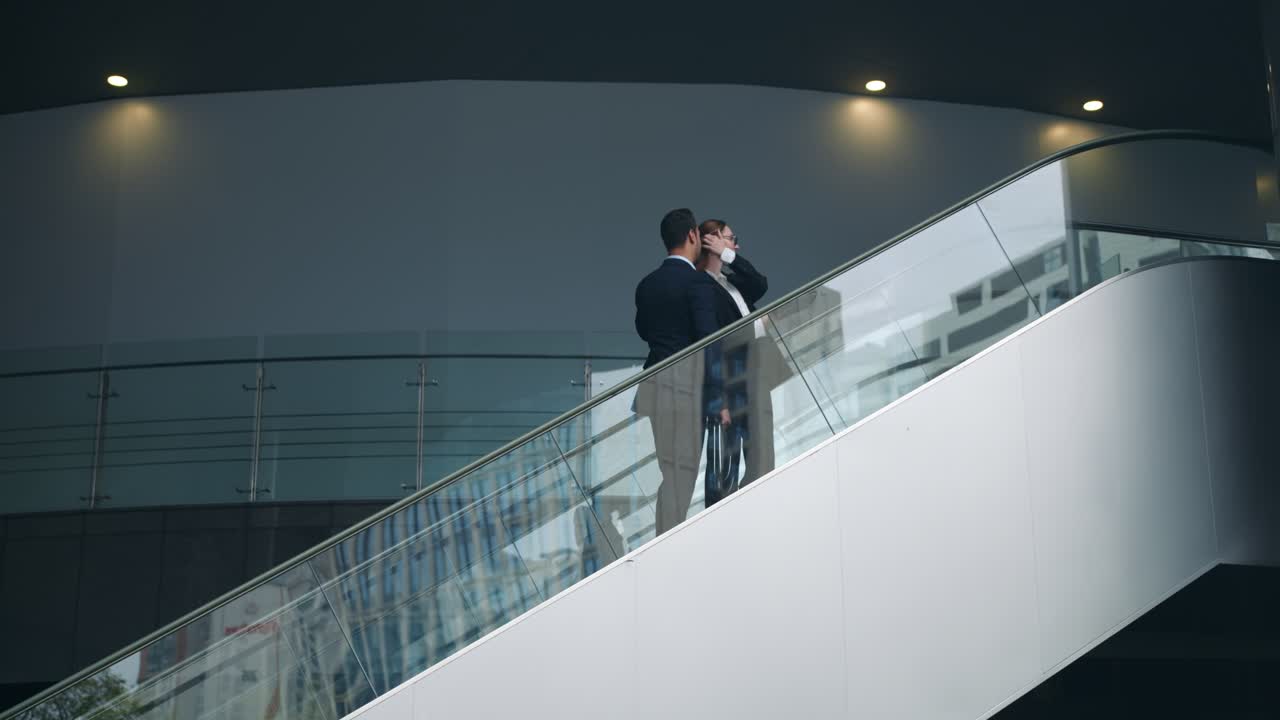 Two smiling business professionals man and woman talk about corporate affairs while ascending on escalator inside contemporary building interior