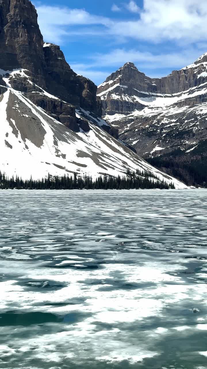 Lago Bow ghiacciato che mostra il ghiaccio che si scioglie, con montagne innevate e cielo blu nel Parco Nazionale di Banff.