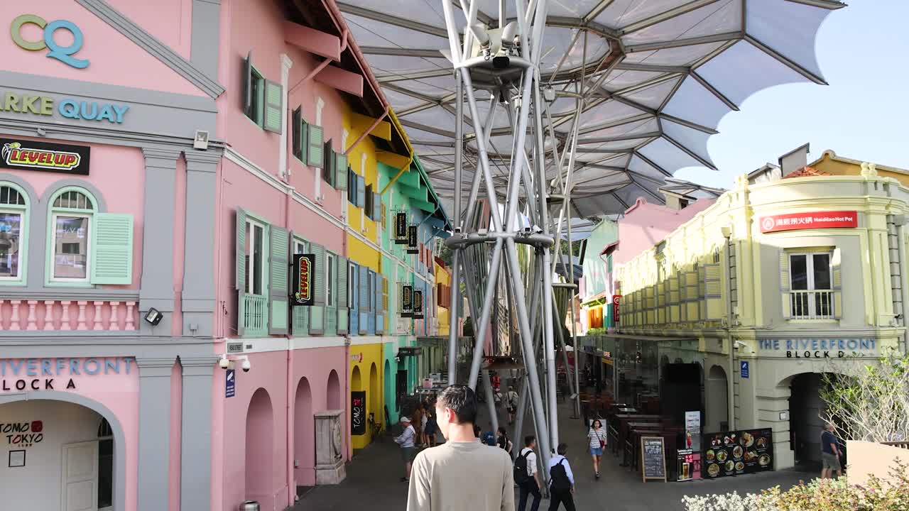 A person walks beneath vibrant pastel shophouses and a modern canopy at Clarke Quay, Singapore, in bright daylight with steady camera movement
