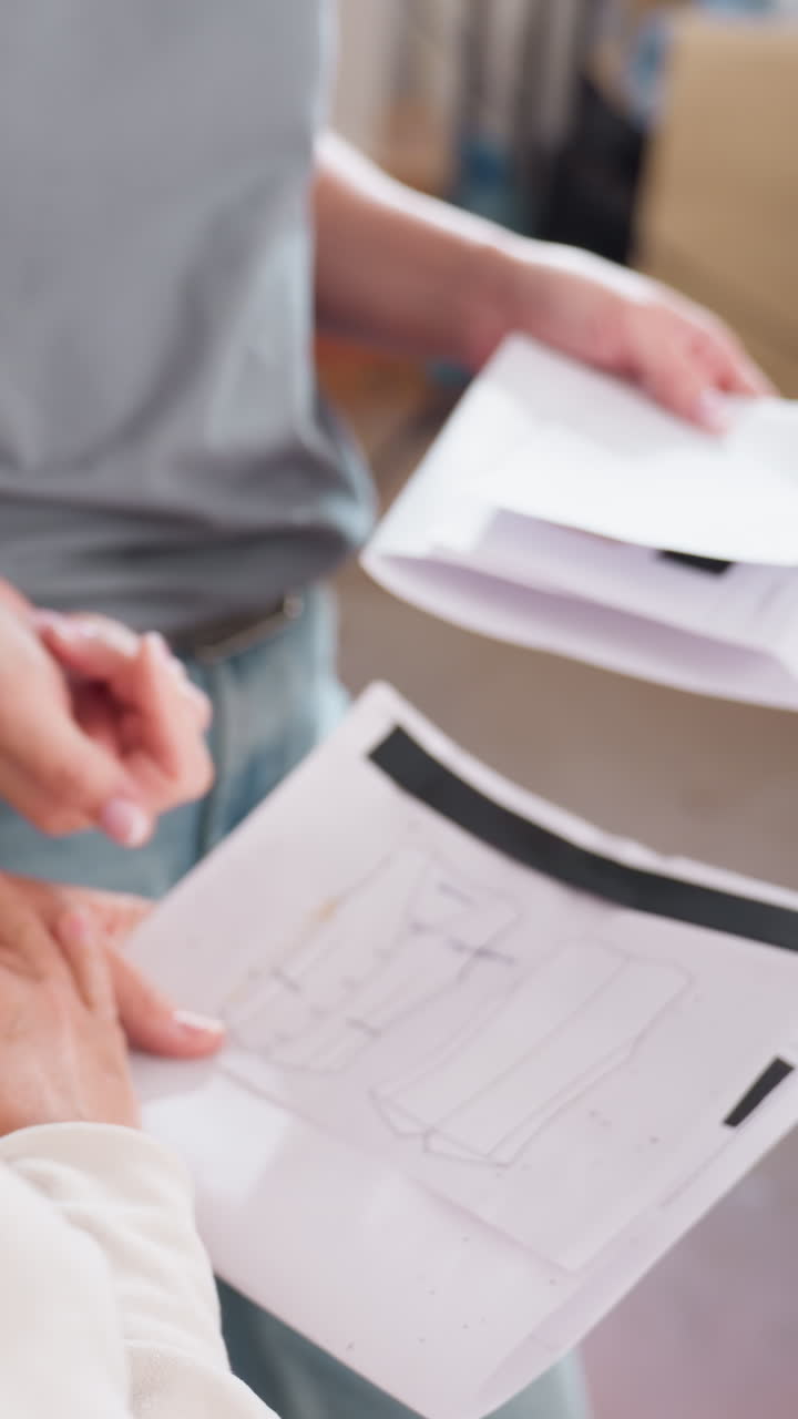 Una mujer blanca pasa hojas de patrones para su aprobación; hay un intercambio de páginas con anotaciones, un lápiz hace las marcas finales; flujo de trabajo de estudio organizado con una pila de patrones y una colaboración centrada.