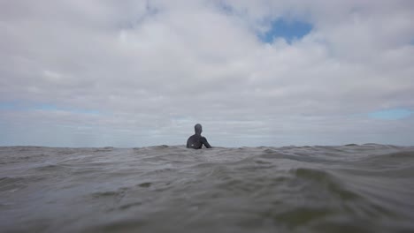 View-along-the-surface-of-sea-water-showing-surfer-in-wet-suit,-floating-in-middle-of-vast-sea