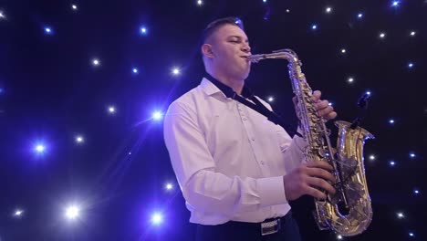 tilt up shot of biracial jazz musician in checkered suit and bowtie playing saxophone while sitting against black band member in dimly lit bar, rehearsing and checking sound before live performance