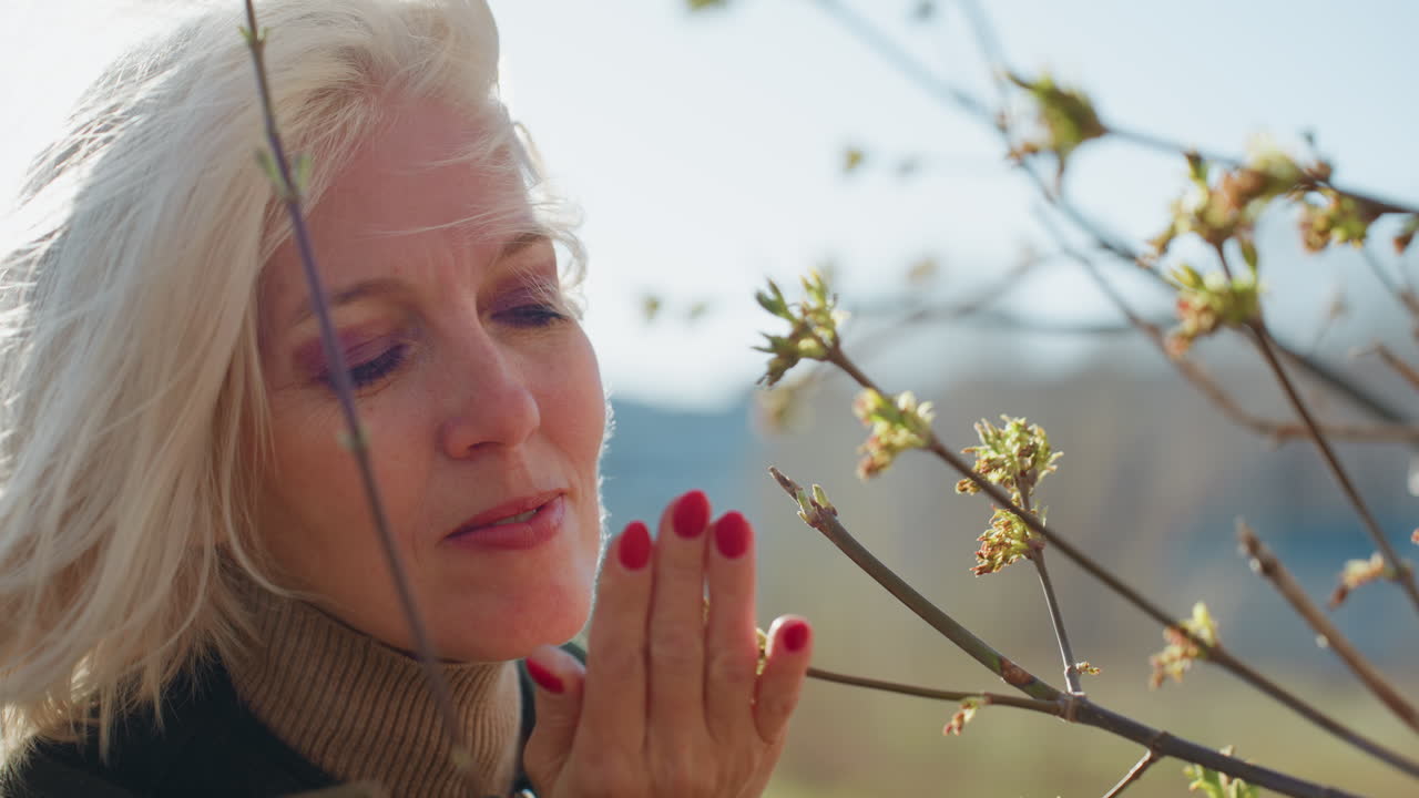 Donna bionda sorridente con occhiali da sole, giacca casual e dolcevita, in piedi all'aperto con le mani giunte vicino ai rami di un albero in fiore in una giornata primaverile soleggiata, che si gode la natura con espressione gioiosa e rilassata