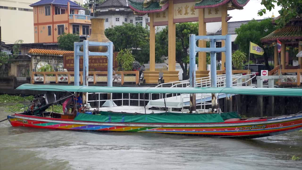 slow motion shot of a boat floating on the Chao Phraya river in Bangkok on a rainy day. Rain drops are visible in the shot. Some traditional construction is on the background.
