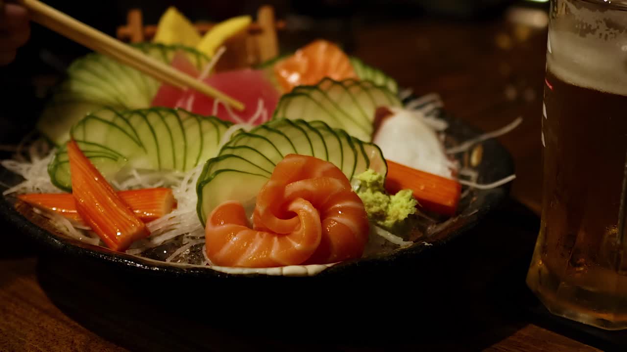 A colorful sashimi platter featuring salmon, cucumber, and tuna paired with a glass of beer on a wooden table.