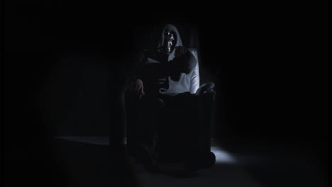 man with scary mask sitting on chair in dark room pointing at camera