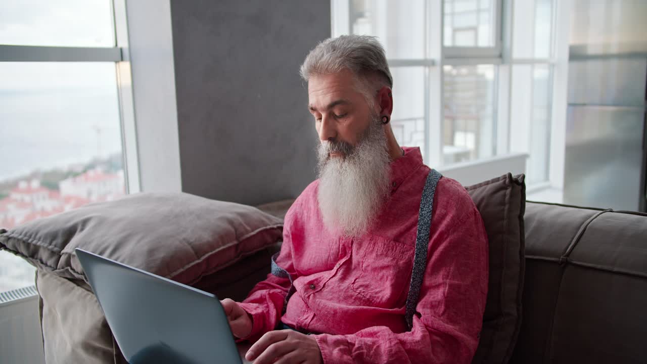 Portrait of an elderly man with gray hair and a lush beard in a pink shirt sits on a brown sofa in a modern apartment and types on a gray laptop
