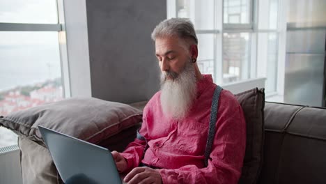 portrait of an elderly man with gray hair and a lush beard in a pink shirt sits on a brown sofa in a modern apartment and types on a gray laptop