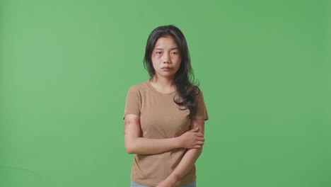 young asian woman with bruise on face and arms hugging herself being hurt from violence while standing on green screen background in the studio