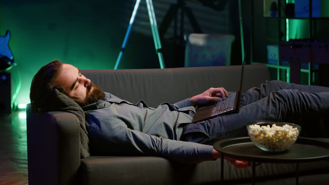 uomo esausto che dorme sul divano dopo aver lavorato sul portatile e visto la tv